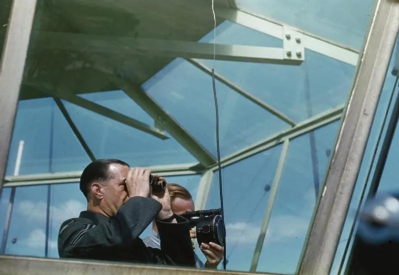 An RAF sergeant looks through binoculars while an assistant aims an Aldis lamp through the angled glazing of the Prestwick tower cab, 1944