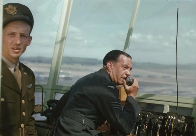 An RAF controller speaks into a hand microphone beside black radio control units on the Prestwick tower desk while a USAAF pilot looks on, 1944