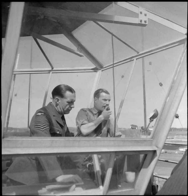 Two controllers at the Prestwick tower desk, one using a hand microphone, with a gooseneck lamp and the airfield beyond the angled glass, 1944